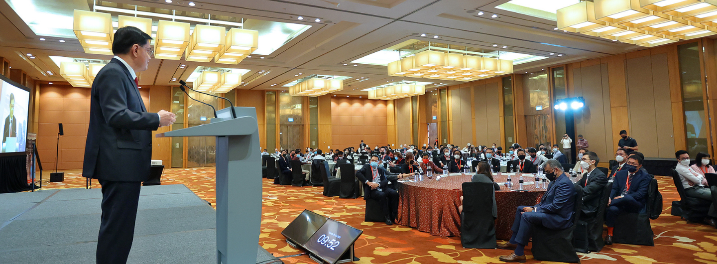Man in suit at podium addresses seated audience in large hall; screen shows his image.